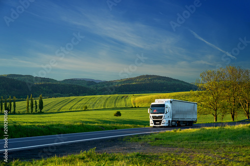 Obraz na płótnie White truck driving on the asphalt road in a rural landscape with forested mountains in the background w salonie