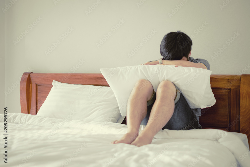 Sad man sitting alone on white bed in bedroom embracing her knees. Worrying about relationships with girlfriend. Vintage filter tone. Sad man sitting alone on white bed in bedroom embracing her knees. Worrying about relationships with girlfriend. Vintage filter tone.