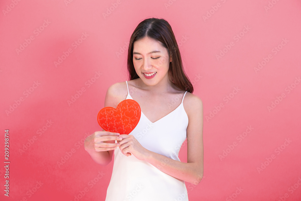 Portrait of attractive Asian smiling woman holding red heart paper jigsaw isolated on pink red background Portrait of attractive Asian smiling woman holding red heart paper jigsaw isolated on pink red background