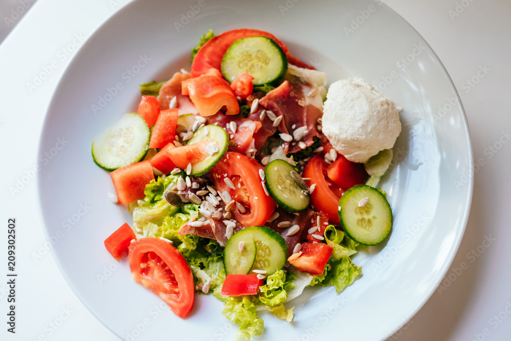 Plate of salad with tomatoes, cucumber, bell pepper, cream cheese, ham on the white table. Glass of iced water with mint and lemon.