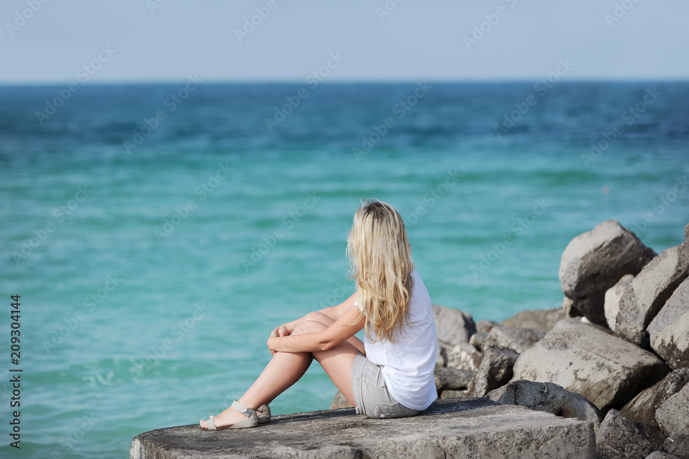 A blonde young woman sitting on a stone with her back looking at the sea