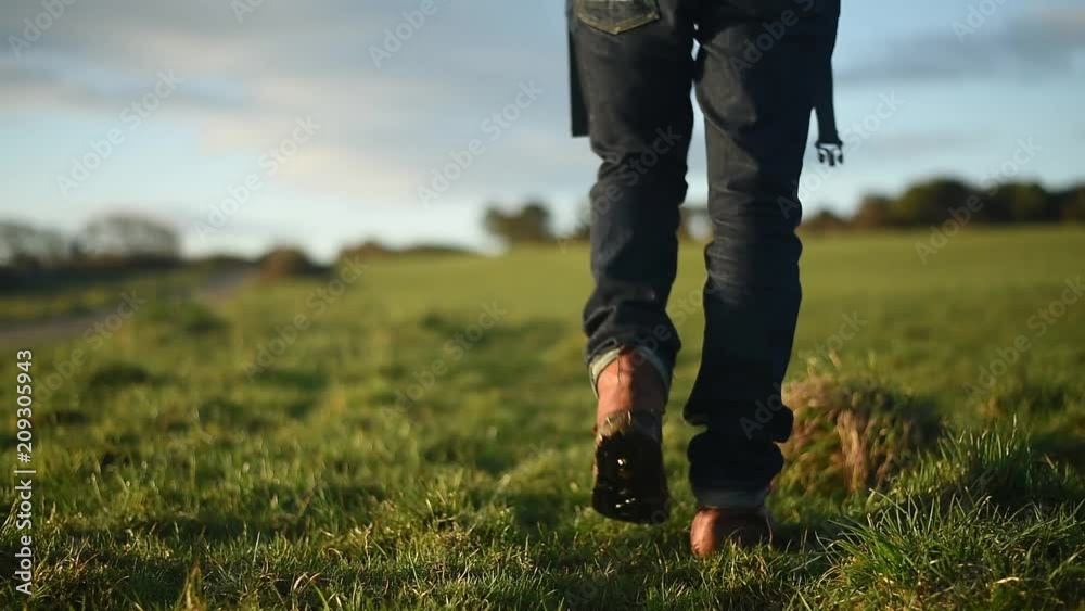 Man in hiking boots and jeans walking across a grass field. Sunrise or sunset. Man in hiking boots and jeans walking across a grass field. Sunrise or sunset.