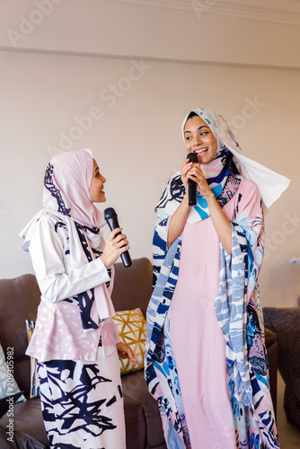 Two young Muslim Malay women unwind and relax as they sing songs on their Karaoke machine at home during the day during Raya. 