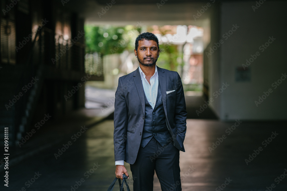 A stylishly dressed Indian Asian man stands in the street in the daytime. He is wearing a 3-piece suit with trendy shoes, a pocket square and earrings. He is holding a gym bag in one hand. A stylishly dressed Indian Asian man stands in the street in the daytime. He is wearing a 3-piece suit with trendy shoes, a pocket square and earrings. He is holding a gym bag in one hand.