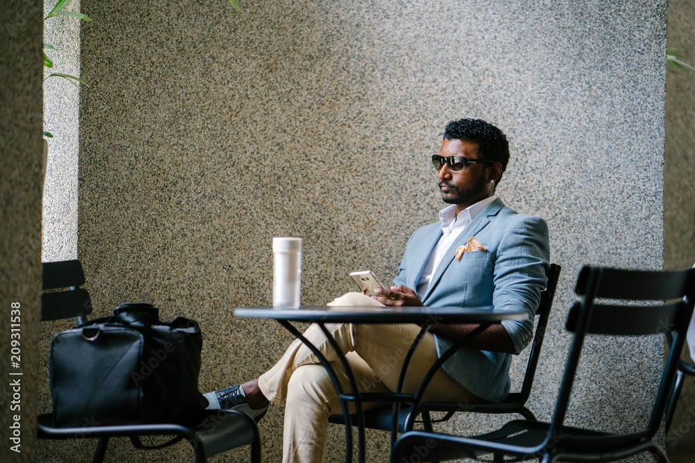 An Indian Asian man is scrolling through his social media feed on his smartphone. He is seated at a co-working space and is wearing a light blue suit.