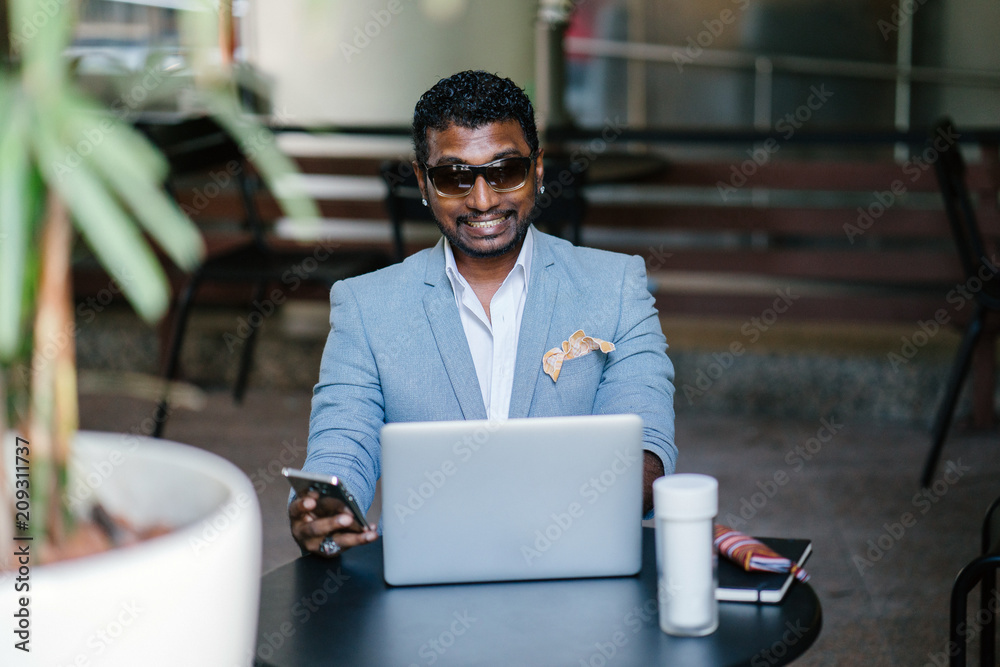 Portrait of an Indian Asian man smiling while checking for new messages on his phone. He is wearing a professionla blue suit and he is working on his laptop in a cafeteria. Portrait of an Indian Asian man smiling while checking for new messages on his phone. He is wearing a professionla blue suit and he is working on his laptop in a cafeteria.