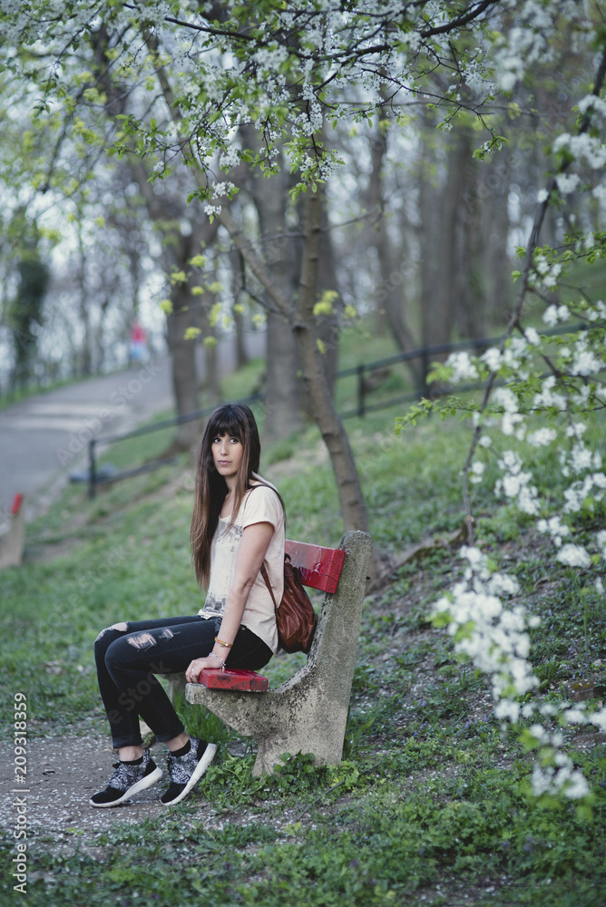 Attractive young woman sitting in a bench outside in park . Caucasian girl with long brown hair in a serene pose. Attractive young woman sitting in a bench outside in park . Caucasian girl with long brown hair in a serene pose.