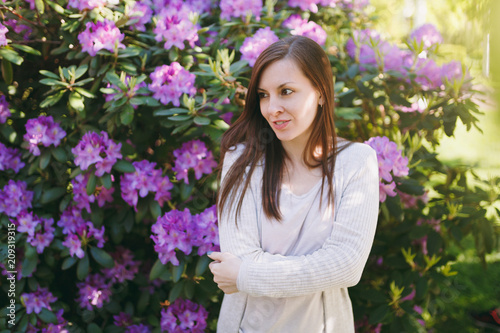 Portrait of beautiful sensual brunette hair young woman with charming smile wearing light casual clothes. Smiling female near flowering bush in city park outdoors on spring nature. Lifestyle concept.