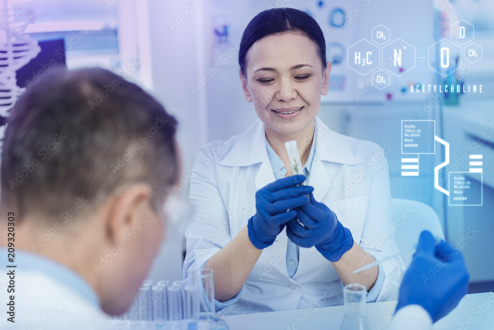 Herb in a test tube. Careful attentive biologist holding a test tube and smiling while looking at the herb in it Herb in a test tube. Careful attentive biologist holding a test tube and smiling while looking at the herb in it