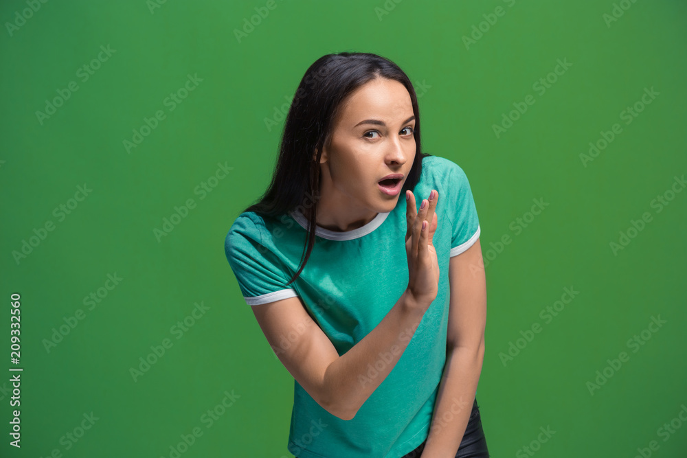 The young woman whispering a secret behind her hand over green background The young woman whispering a secret behind her hand over green background
