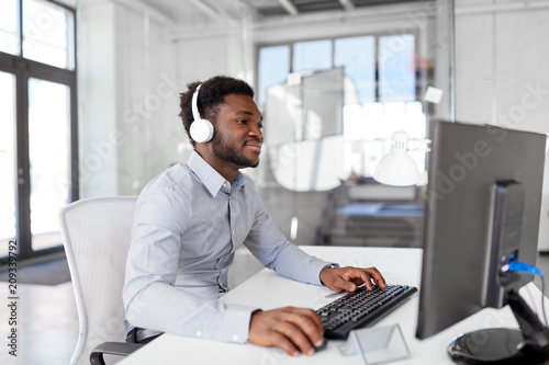 business, technology and people concept - happy african american businessman with headphones and computer listening to music at office