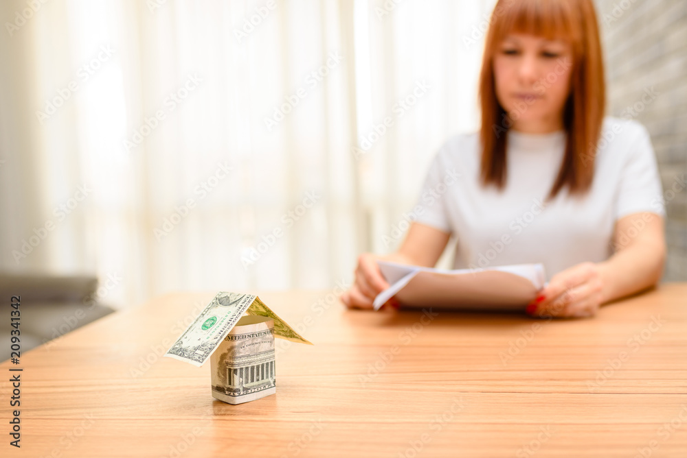 Money house from dollar banknote on wooden table.Happy woman sitting and calculating bills in the home office. Concept of buying a home, real estate activity, looking at new houses for sale. Money house from dollar banknote on wooden table.Happy woman sitting and calculating bills in the home office. Concept of buying a home, real estate activity, looking at new houses for sale.