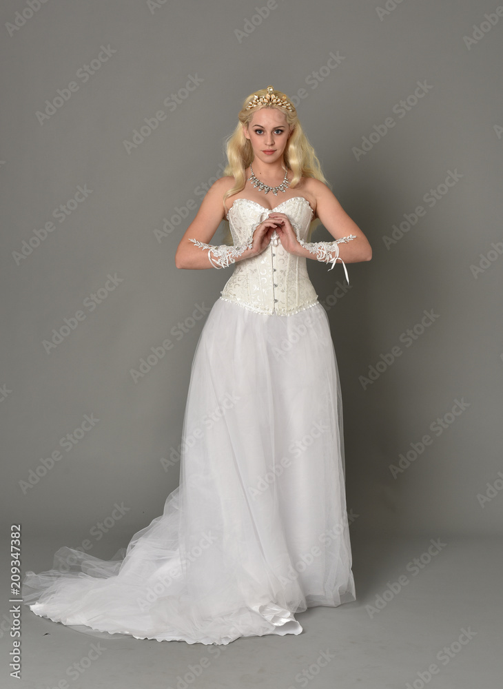 full length portrait of blonde girl wearing white corset gown. standing pose on grey studio background.