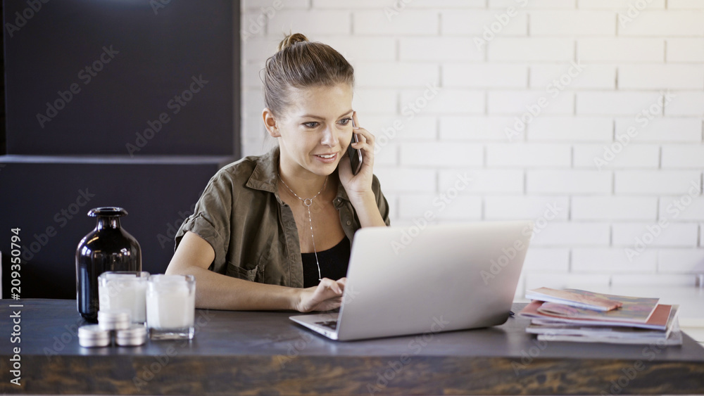 Fototapeta premium A young busy smiling blond casual pretty woman freelancer businesswoman is working at home with a laptop having a call background of a white brick wall.