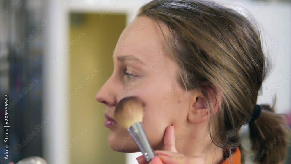 Side view of a young, pretty female model. Make up process. Artist putting some transparent, shiny rouge on woman's cheek using a brush Side view of a young, pretty female model. Make up process. Artist putting some transparent, shiny rouge on woman's cheek using a brush