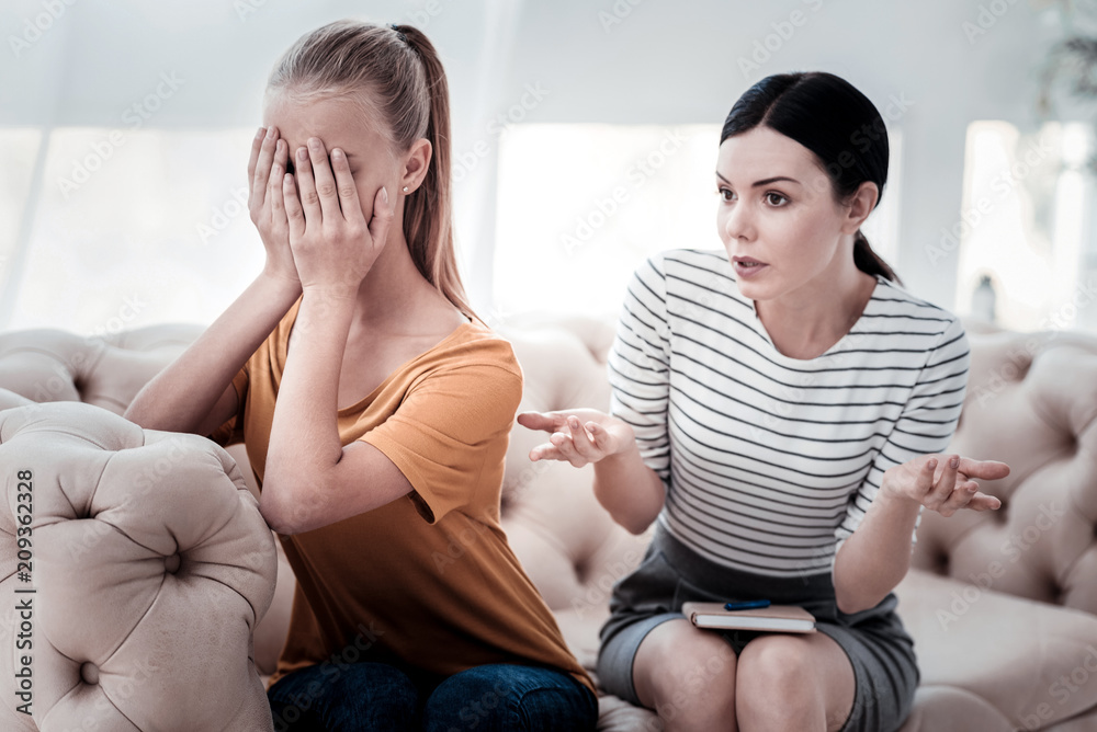 Behavior problems. Portrait of crying girl covering her face with hands while skilled psychologist having expressive talk with her Behavior problems. Portrait of crying girl covering her face with hands while skilled psychologist having expressive talk with her