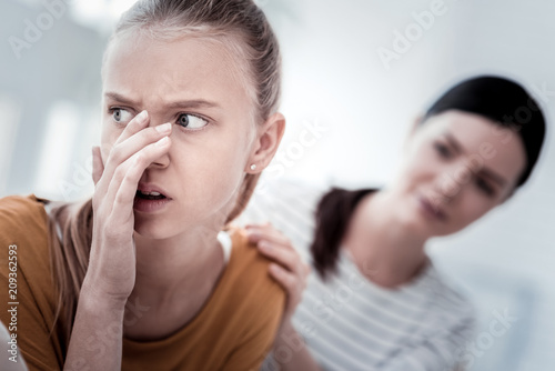 Unstable moral world. Close up of frustrated girl covering her face with a hand and looking away and her mother standing next to her and putting her hand on girls shoulder