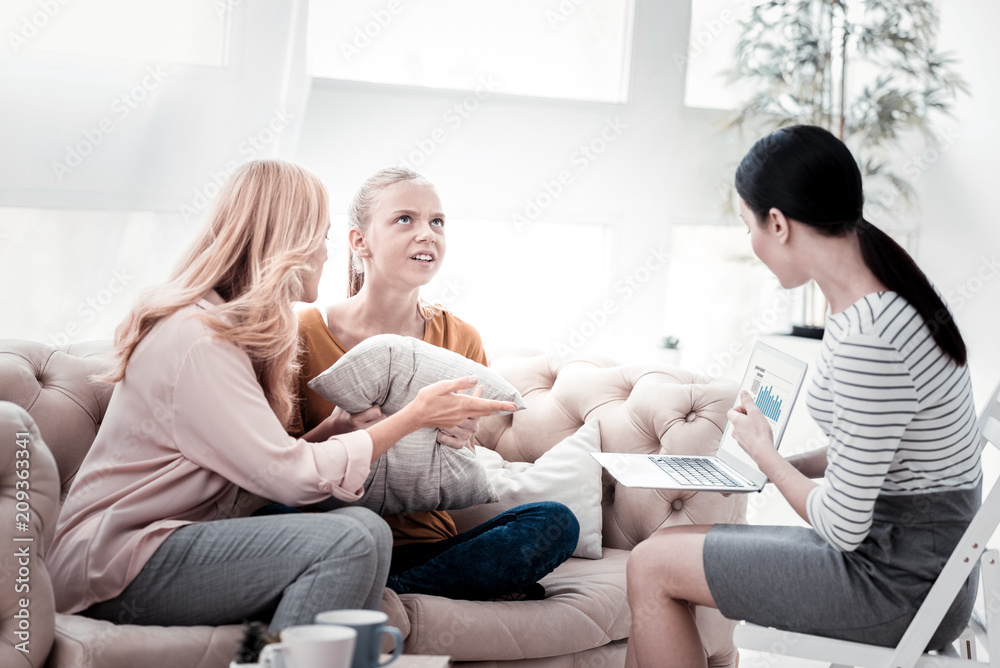 Wrong information. Irritated girl holding pillows and expressing negativity while her mother and psychologist using a laptop and pointing at her problems Wrong information. Irritated girl holding pillows and expressing negativity while her mother and psychologist using a laptop and pointing at her problems