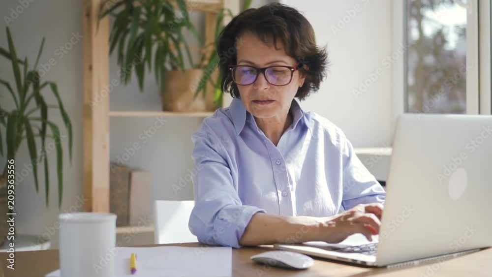 Elderly businesswoman is working with laptop sitting in home office space, successful mature lady wearing glasses is doing job using PC at table in room with daylight. Concept: business person, modern Elderly businesswoman is working with laptop sitting in home office space, successful mature lady wearing glasses is doing job using PC at table in room with daylight. Concept: business person, modern