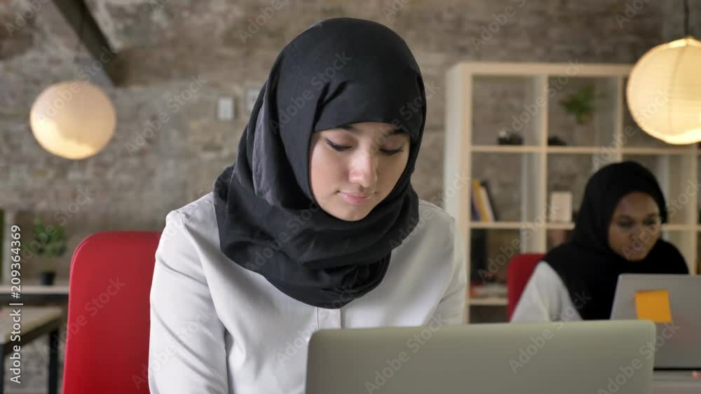 Portrait of young muslim women in hijab working and looking in camera, smiling, two womens sitting in modern office