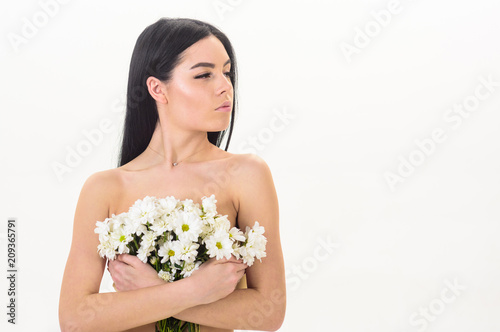 Girl on calm face stands naked and holds chamomile flowers in front of chest. Lady covers breasts with flowers, isolated on white. Skin health concept. Woman with smooth healthy skin looks attractive.