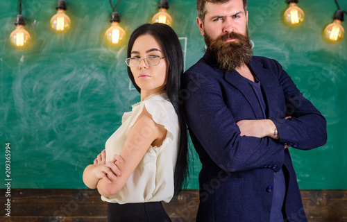 Man with beard and teacher in eyeglasses stand back to back, chalkboard on background. Teacher and schoolmaster look confident. School staff concept. Lady and hipster working together in school.