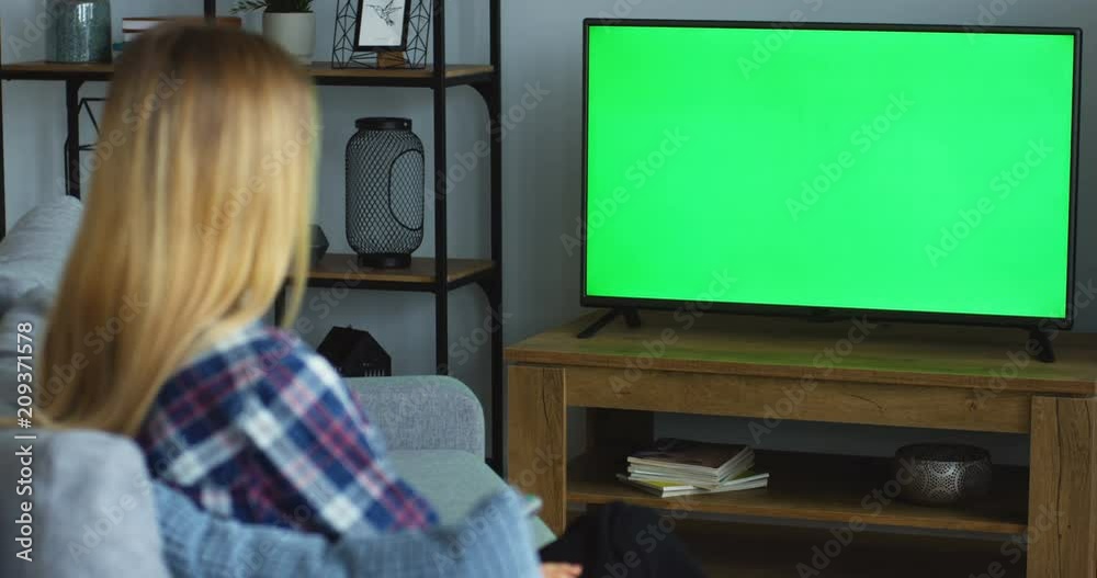 Rear of the blonde woman in the motley blue shirt sitting on the gray couch in the living room, watching TV with green screen and changing channels with a remote control. Chroma key. Inside