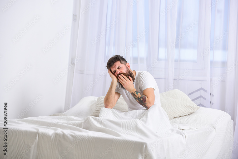 Guy on sleepy tired face yawning. Macho with beard and mustache yawning, relaxing, having nap, rest. Man in shirt sits on bed, white curtains on background. Nap and siesta concept. Guy on sleepy tired face yawning. Macho with beard and mustache yawning, relaxing, having nap, rest. Man in shirt sits on bed, white curtains on background. Nap and siesta concept.