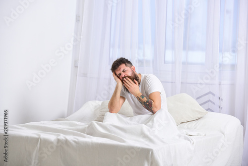 Guy on sleepy tired face yawning. Macho with beard and mustache yawning, relaxing, having nap, rest. Man in shirt sits on bed, white curtains on background. Nap and siesta concept.