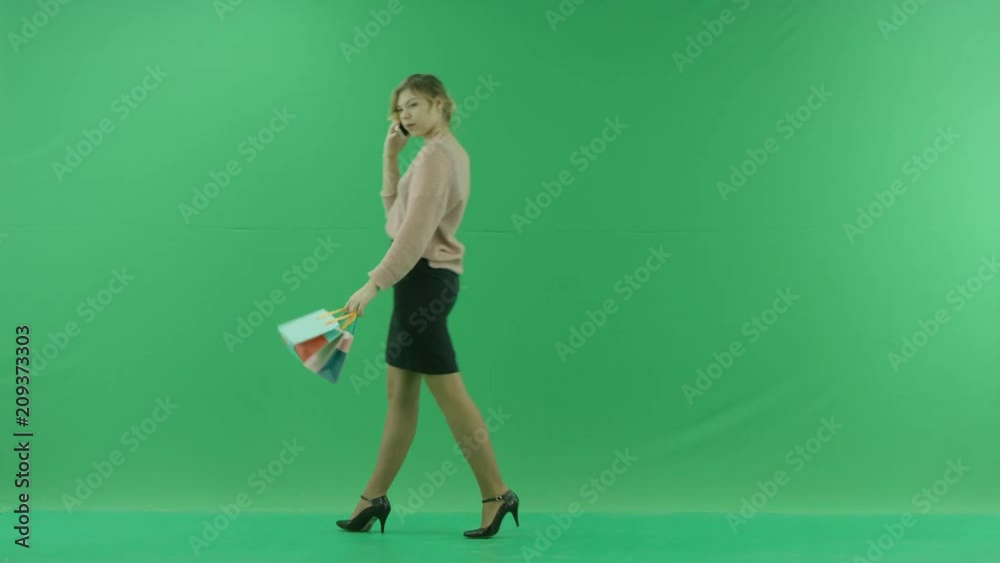 Pretty shopping woman is walking with phone and shopping bags. Female shopper holding shopping bags sideways on green background in studio. Pretty shopping woman is walking with phone and shopping bags. Female shopper holding shopping bags sideways on green background in studio.