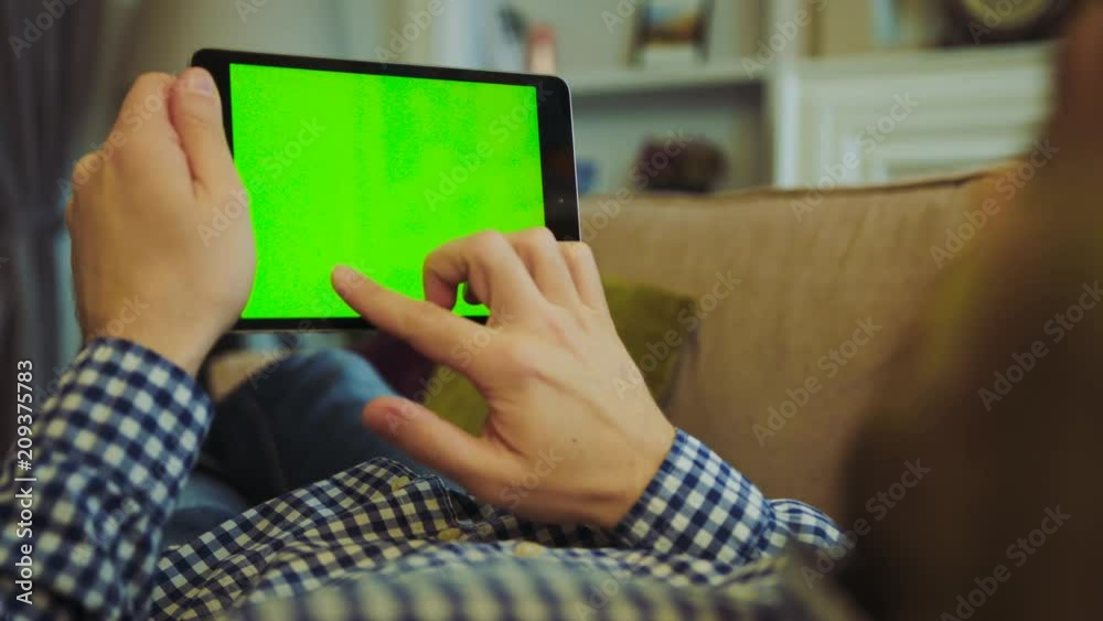 Man in plaid shirt and jeans lying on the cozy sofa and scrolling on the green screen of black tablet device in his hands. Chroma key. View over the shoulder. Close up. Resting at home. Indoors Man in plaid shirt and jeans lying on the cozy sofa and scrolling on the green screen of black tablet device in his hands. Chroma key. View over the shoulder. Close up. Resting at home. Indoors