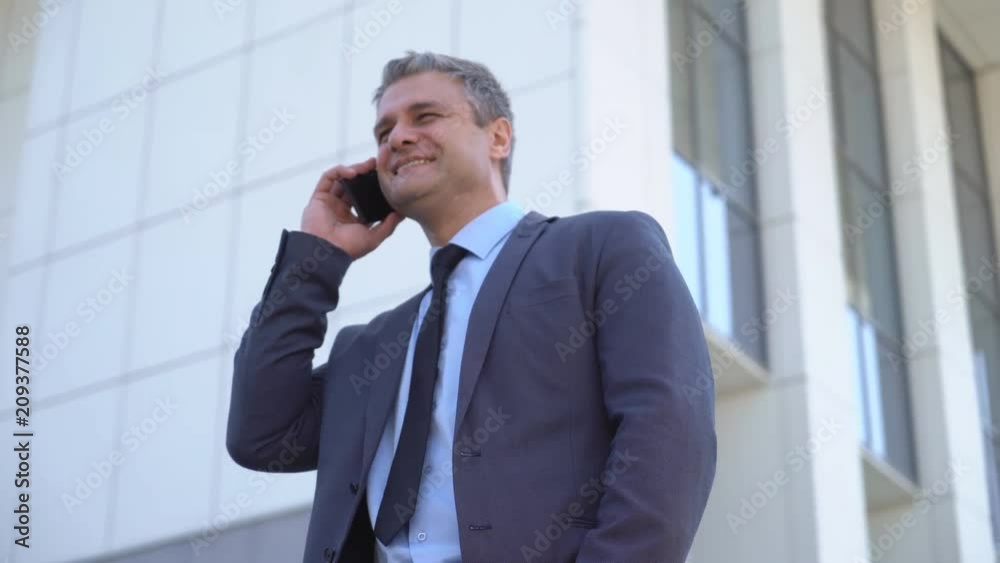 A gray-haired middle-aged man in a suit is looking at the camera against the backdrop of a business center. A man is talking on the phone. Business and success. portrait to the waist 4 к A gray-haired middle-aged man in a suit is looking at the camera against the backdrop of a business center. A man is talking on the phone. Business and success. portrait to the waist 4 к