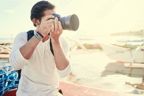 Photography and travel. Young man with rucksack taking photo with his camera enjoying beautiful tropical sea view on fishing beach.
