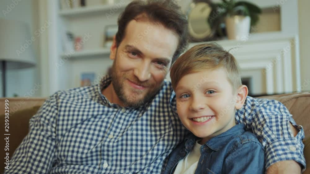 Portrait shot of the smiling happy father with beard abd cute little son sitting on the couch in the living room. Close up. Posing. Indoors Portrait shot of the smiling happy father with beard abd cute little son sitting on the couch in the living room. Close up. Posing. Indoors