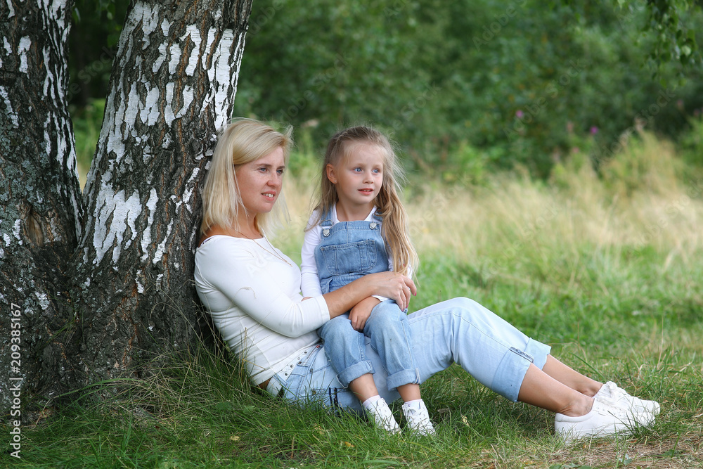 Happy family in nature. Beautiful and happy mother gently hugs her little daughter with blond hair against the background of grass while walking in the summer park. Happy Family Outdoor. Happy family in nature. Beautiful and happy mother gently hugs her little daughter with blond hair against the background of grass while walking in the summer park. Happy Family Outdoor.