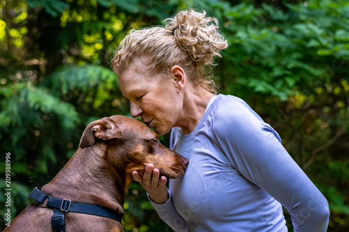 Female blond baby boomer in a purple top in a quiet loving moment with her two tone brown rescue dog out in the woods
 photo
