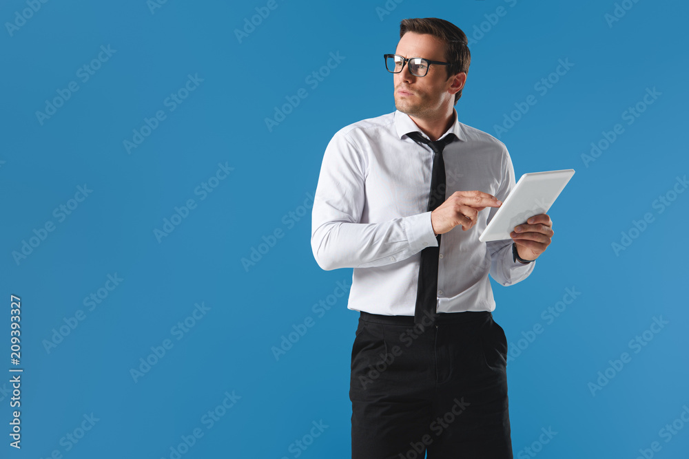 serious businessman in eyeglasses using digital tablet and looking away isolated on blue