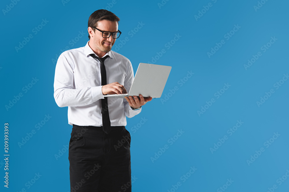 smiling handsome businessman in eyeglasses using laptop isolated on blue smiling handsome businessman in eyeglasses using laptop isolated on blue