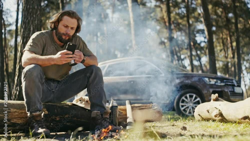 Muscular casual man sitting with axe at burning campfire in forest and using phone on background of car.