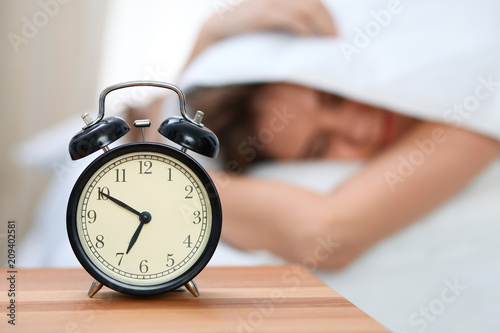 Sleepy young woman under blanket with her head don't want to wake up in front of alarm clock closeup. Early wake up, not getting enough sleep concept