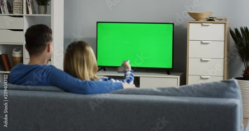Rear of the romantic couple hugging on the couch while watching TV with green screen and changing channels in the living room. Indoors