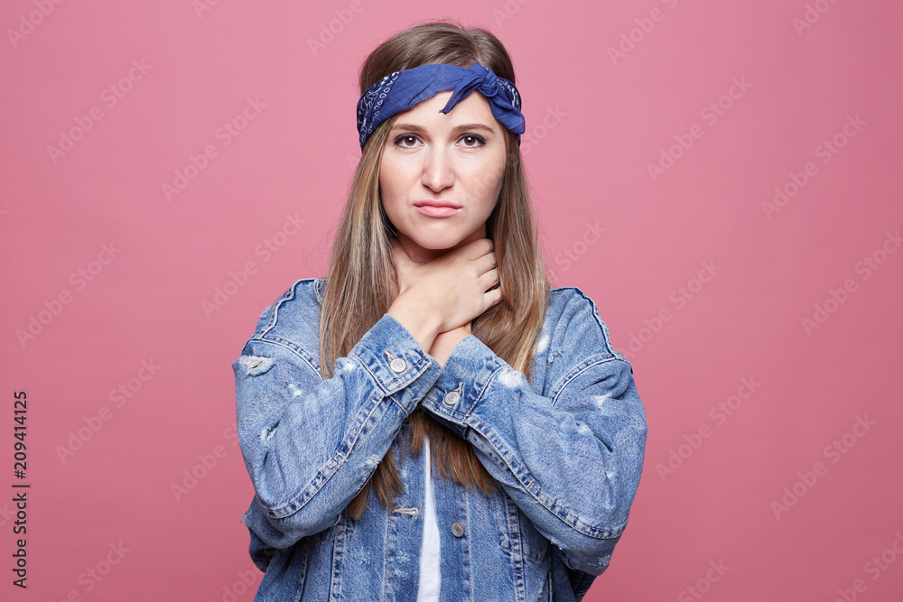 Attractive female in despair makes suicide gestures and smothers herself, suffers from suffocation, wears oversized denim and bandana, poses against pink background. People, body language concept