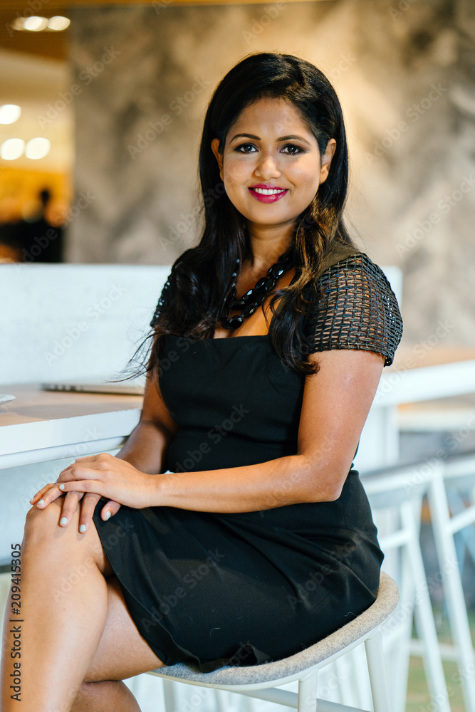 Portrait of a confident, successful and attractive Indian Asian business woman sitting on a chair in a corridor of her office in the day. She is smiling and looking very classy for her professional he