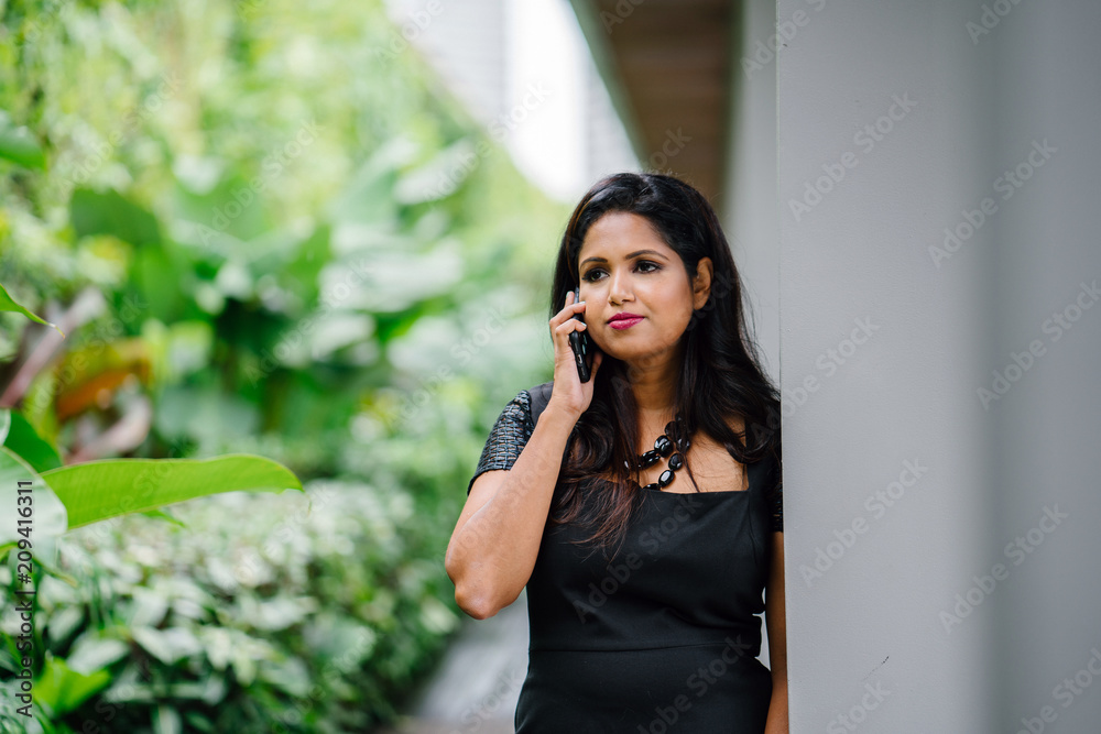 A young desi Asian woman is talking on the phone with her business partner. She looks serious and is standing beside green plants. A young desi Asian woman is talking on the phone with her business partner. She looks serious and is standing beside green plants.