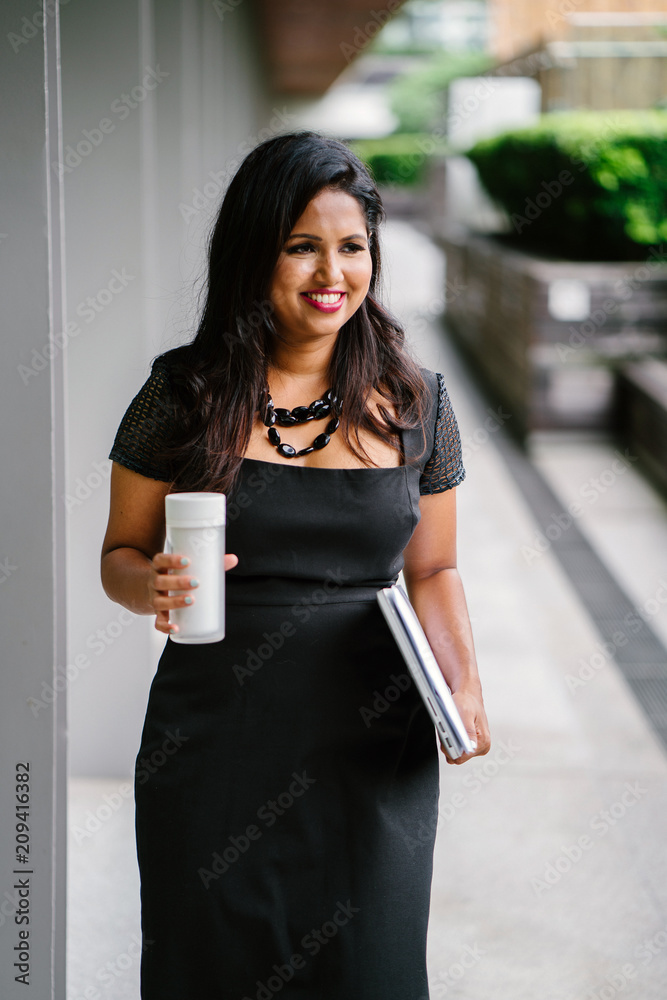 A young Indian Asian business woman grabs some coffee as she heads to her next client meeting. She looks happy as she walks along a shaded path. A young Indian Asian business woman grabs some coffee as she heads to her next client meeting. She looks happy as she walks along a shaded path.