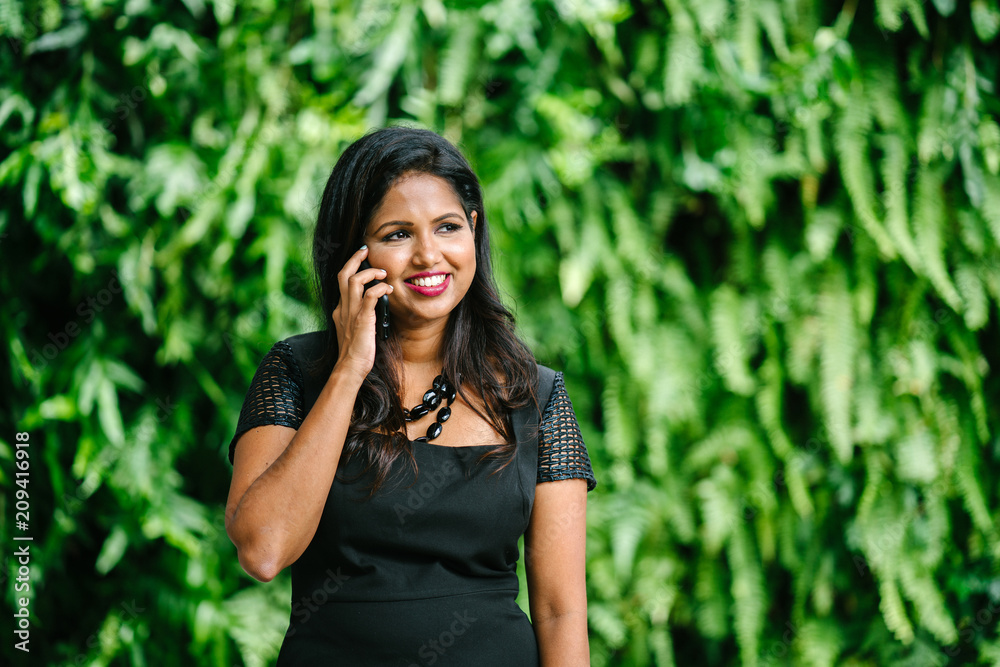 A young Indian Asian woman is speaking to her colleague using her smartphone. She is standing in front of a green wall of plants and is wearing a black dress. A young Indian Asian woman is speaking to her colleague using her smartphone. She is standing in front of a green wall of plants and is wearing a black dress.