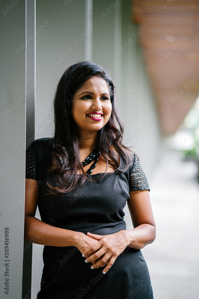 An Indian Asian professional woman leaning on a pillar while standing along the corridor on a sunny day. She is smiling confidently and looks dignified in her black dress. An Indian Asian professional woman leaning on a pillar while standing along the corridor on a sunny day. She is smiling confidently and looks dignified in her black dress.