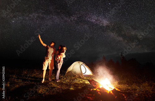 Tourist family camping in mountains at night. Mother holding in arms small daughter, father pointing at bright stars in dark sky and Milky way in front of illuminated tent and burning bonfire