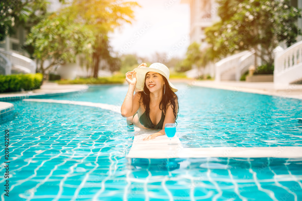 Asian woman in bikini and hat, enjoying cocktail in a swimming pool, summer time