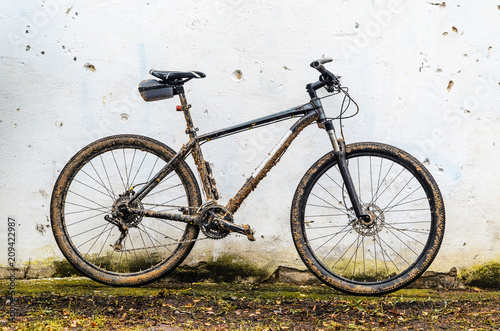 Dirty Mountain Bike Covered with Mud After Riding in Bad Weather Stands. Grey 29er Hardtail Bike on White Wall Background